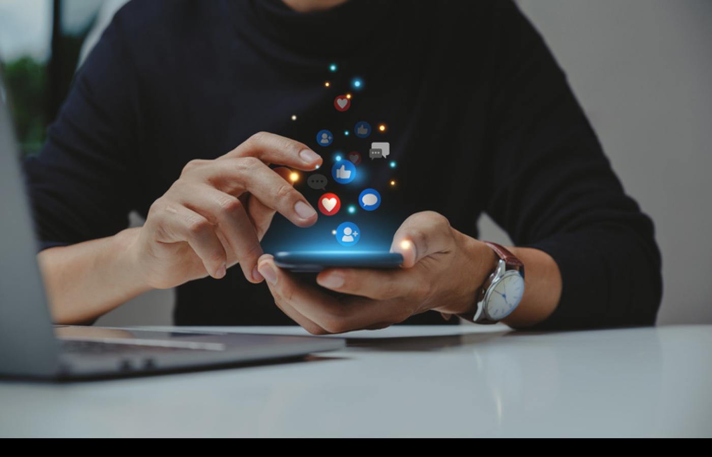 Hands tapping on a mobile phone with social media icons glowing above the phone.